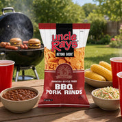 A bag of Uncle Ray's BBQ Pork Rinds chips stands on a wooden table outdoors, surrounded by red plastic cups, bowls of food, and a grill in the background.