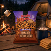 A bag of Uncle Rays Cinnamon Brown Sugar Mini Pretzels stands on a wooden camp table at night, surrounded by a campfire, a mug of hot cocoa with marshmallows, graham crackers, and a lantern, with a tent in the background.