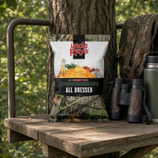 A bag of Uncle Rays Money Oak Competition All Dressed potato chips is displayed on a wooden hunting stand next to a tree trunk, with a camouflage backpack, binoculars, and a thermos in a forest setting.