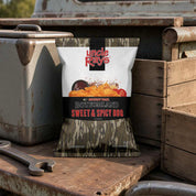 A bag of Uncle Ray's Bottomland Sweet & Spicy BBQ Flavored Potato Chips stands on a weathered wooden surface next to a metal toolbox and wooden crates. The packaging features a white top half with the red and black Uncle Ray's logo, a central image of golden-brown chips with red pepper flakes, and a camouflage-patterned bottom half.