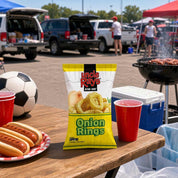 A bag of Uncle Rays Onion Rings snack stands on a wooden table at an outdoor barbecue or tailgate, surrounded by red plastic cups, hot dogs, a soccer ball, and a grill in a parking lot setting.