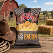 A bag of Uncle Rays Sweet Black Pepper Bacon flavored potato chips stands on a wooden table outdoors, with a red barn, fence, hay bales, a brown cowboy hat, and rope in the background. The bag features bacon graphics and bold red and yellow text.